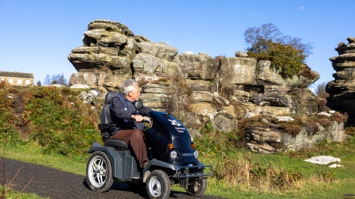Visitor using a mobility scooter on an accessible tarmac path at Brimham Rocks, with large weathered rock formations and autumn vegetation in the background under a clear blue sky.